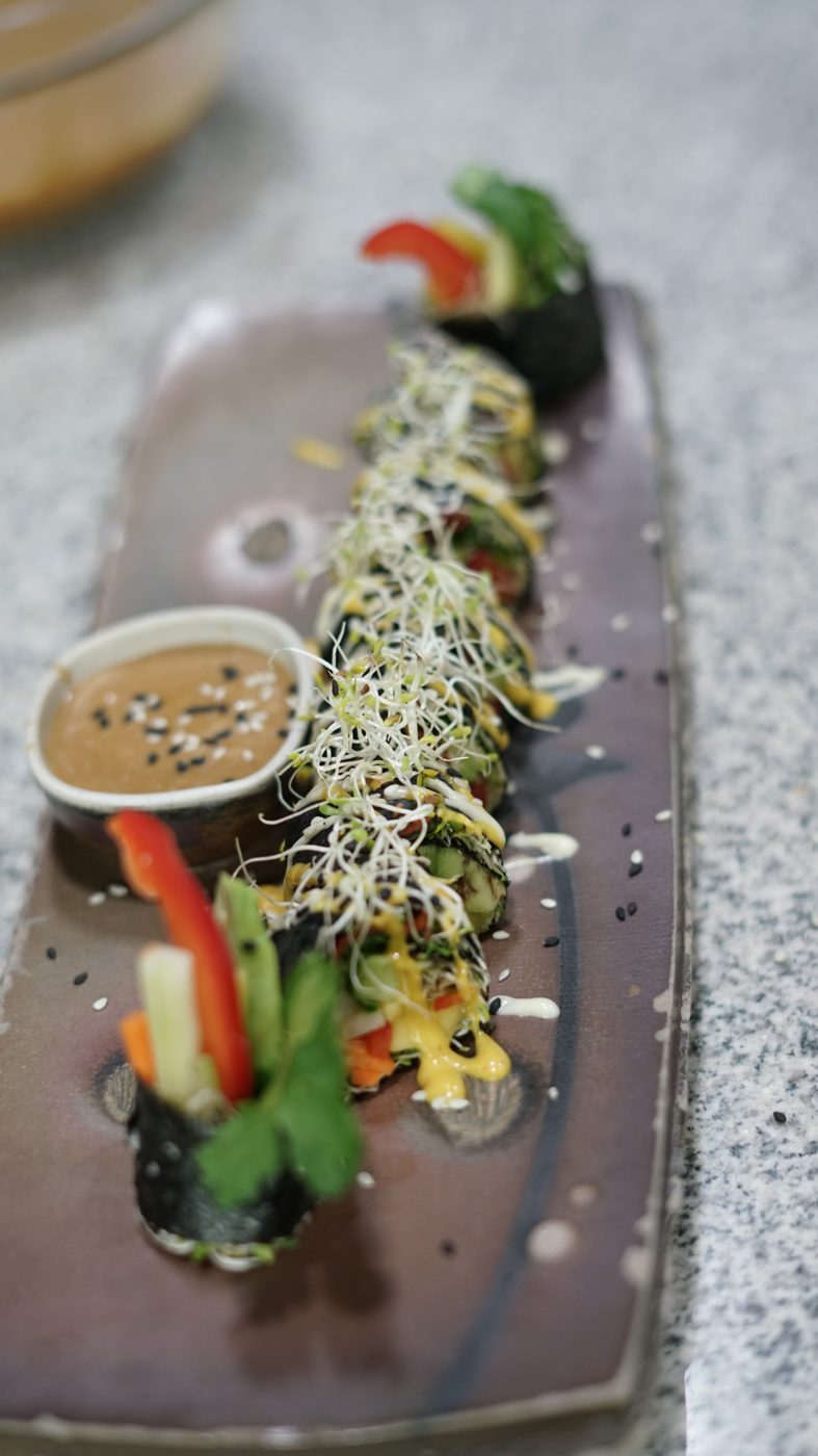 A sliced vegetable sushi roll with sprouts and dipping sauce on a stone plate